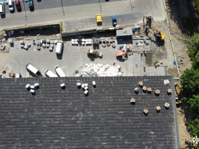 Widok z drona nad budynkiem sklepu w Ustrzykach Dolnych, widać dach na nim materiały do pokrycia dachu. Z przodu sklepu widać parking, pracownicy jeszcze go budują. Samochody dostawcze zaparkowane i materiały budowlane obok.