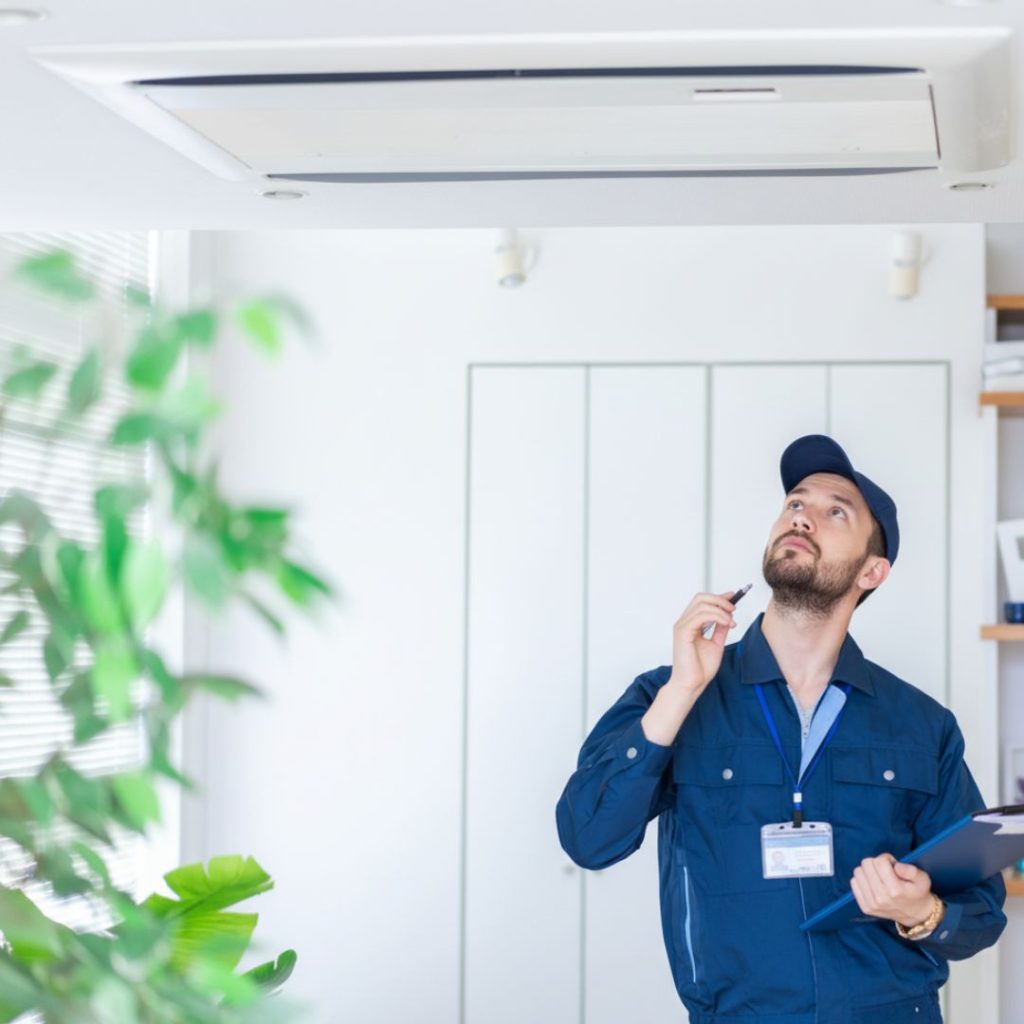 Mężczyzna technik w niebieskim uniformie z czapką i brodą, stojący w jasnym pomieszczeniu biurowym, patrzy w górę na zamontowaną na suficie jednostkę klimatyzacyjną. Trzyma długopis i clipboard, sprawdzając urządzenie.